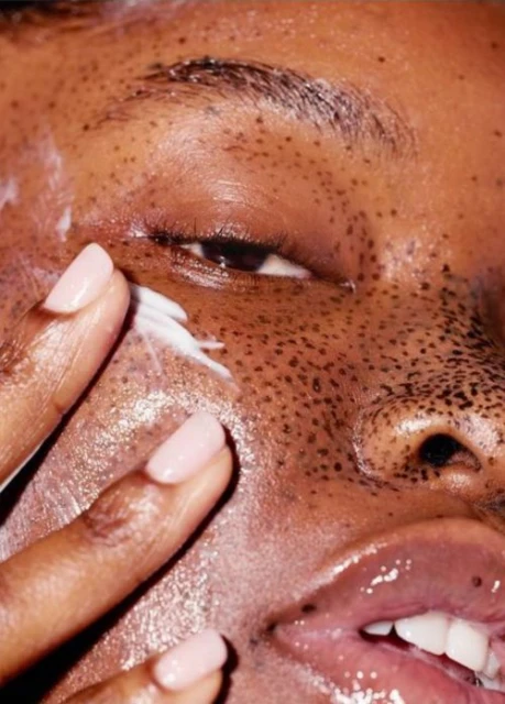 Close-up of a woman applying white skincare cream to her face, with her eye, fingers, and glossy lips in sharp focus.