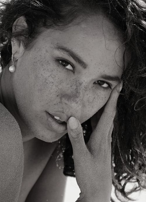 Close-up black and white portrait of a freckled woman with dark curly hair touching her face, captured by Ali Mitton.