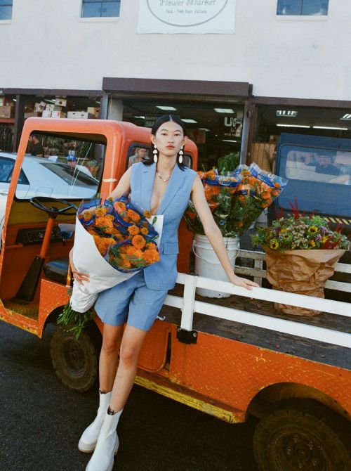 Woman in blue suit poses with flowers near orange utility vehicle outside a flower market on a sunny day. White ankle boots.