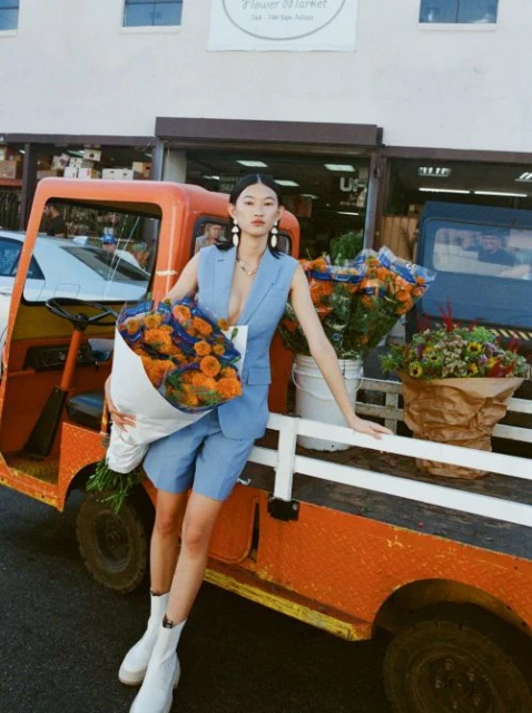 Woman in blue suit poses with flowers near orange utility vehicle outside a flower market on a sunny day. White ankle boots.