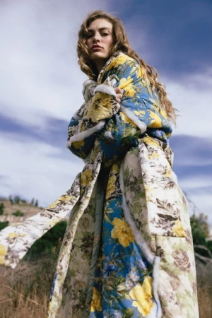 Model with long wavy hair wearing a blue and yellow floral quilted coat outdoors under an overcast sky.