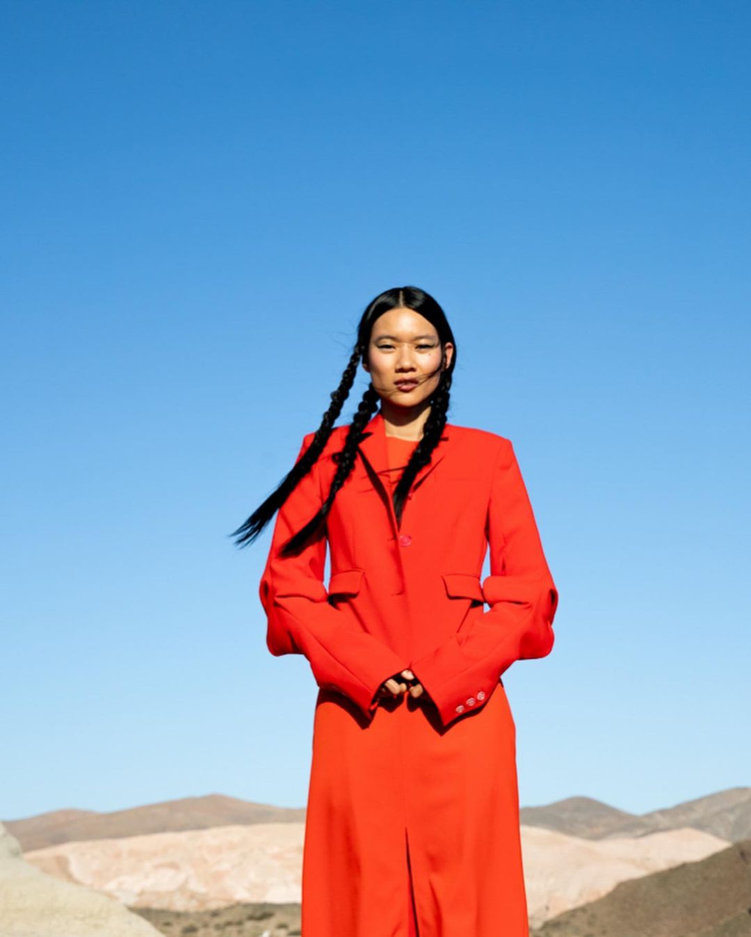 Young woman in red blazer and dress poses in desert landscape under blue sky. Braided hair and hands clasped in fashion portrait.