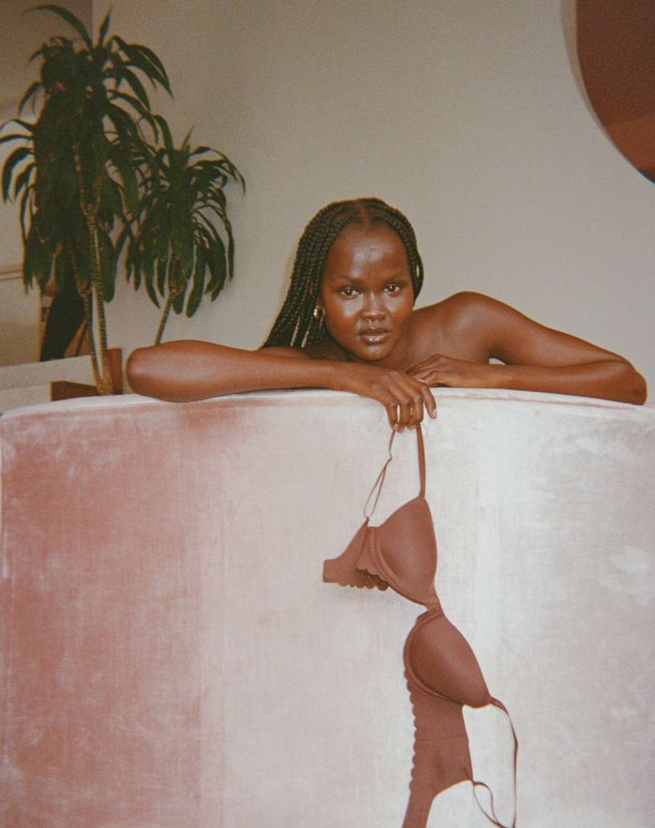 Adult female poses with a bra in an indoor setting. She leans against a tub with a plant in the background.