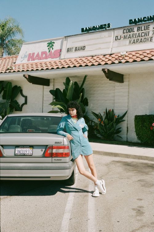 Young woman in turquoise dress leans against a silver Volvo car outside a banquet hall with palm trees on a sunny day.