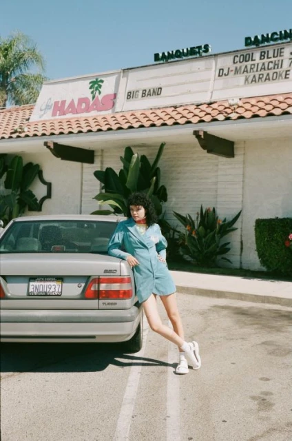 Young woman in turquoise dress leans against a silver Volvo car outside a banquet hall with palm trees on a sunny day.