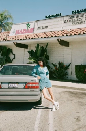 Young woman in turquoise dress leans against a silver Volvo car outside a banquet hall with palm trees on a sunny day.