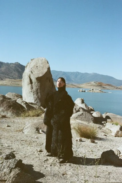 An Asian woman in a dark coat poses near a large rock formation by a lake under a clear sky, creating a scenic outdoor fashion shot.