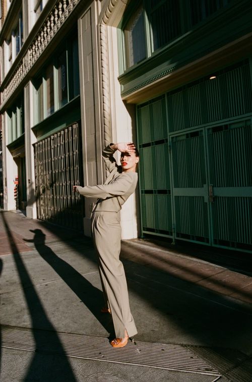 Woman in stylish tan suit posing outdoors in urban setting with hand shielding eyes. Sunlight creates strong shadows on street and building.