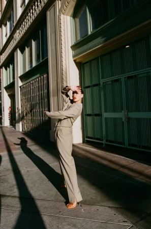 Woman in stylish tan suit posing outdoors in urban setting with hand shielding eyes. Sunlight creates strong shadows on street and building.