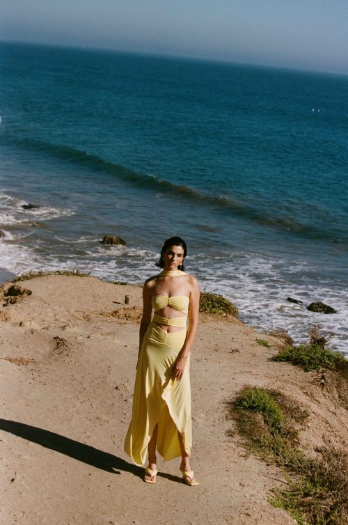 A woman in a yellow dress poses on a cliff overlooking the ocean under a clear sky. The sunlight creates a relaxed atmosphere.