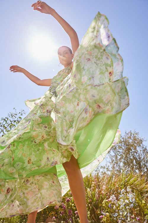 Woman in flowing floral dress with arms raised stands outdoors against a blue sky backdrop with trees and natural light creating lens flare.