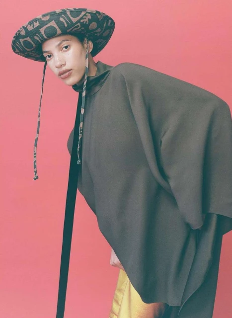 A young woman in a flowing black top and patterned hat poses against a coral background, showcasing contemporary fashion and style.