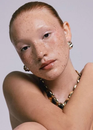 Close-up portrait of a woman with heavy freckles wearing gold hoop earrings and a statement necklace against a plain grey background.