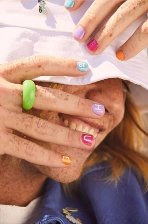 Close-up portrait of a smiling person wearing a white bucket hat, bright colorful nail polish, and a chunky green resin ring.