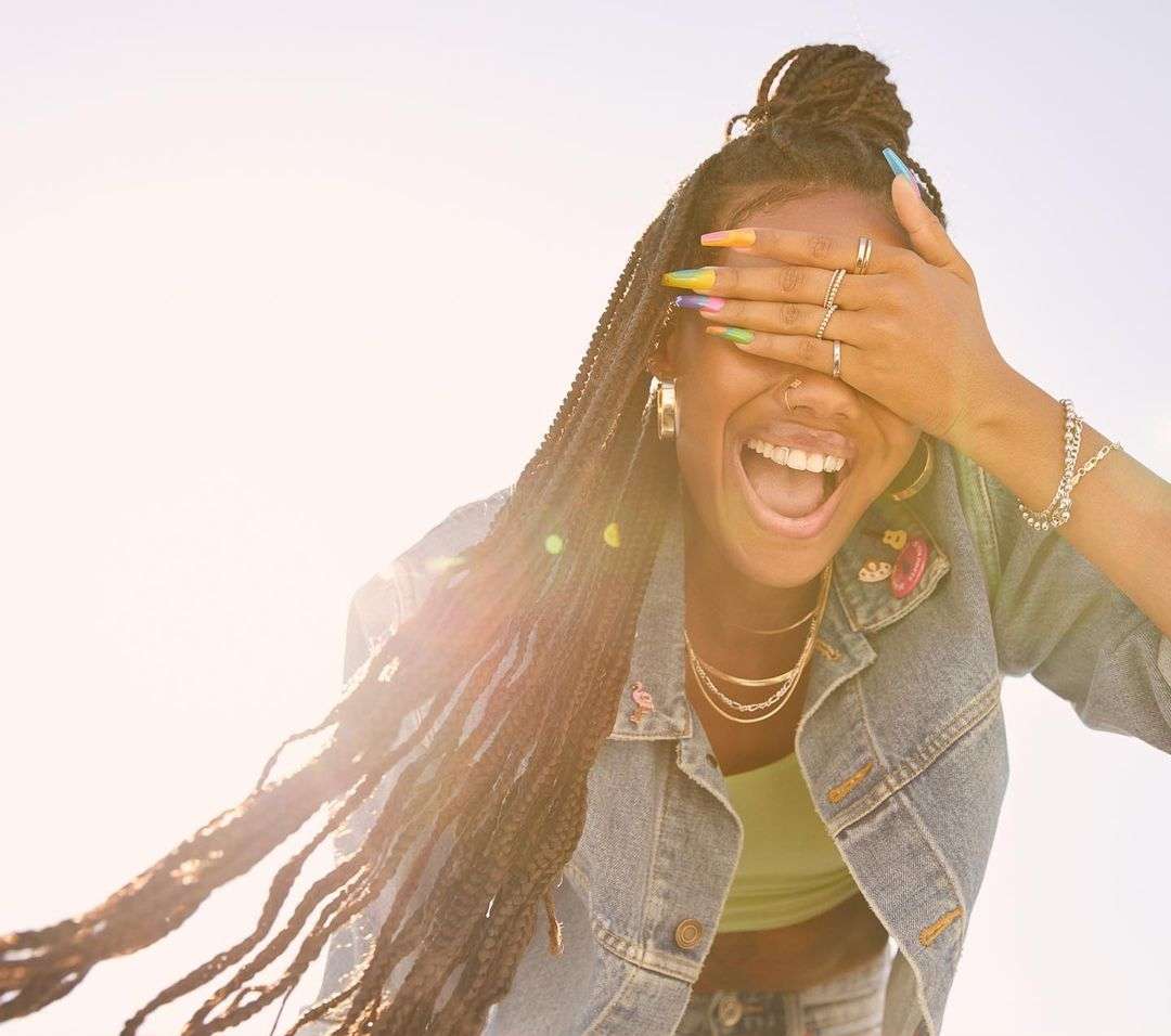 Smiling woman with braids covers her eyes with manicured nails, wearing a denim jacket outdoors on a sunny day, posing cheerfully.