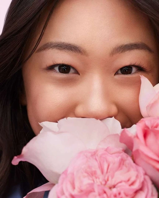 Close-up of a young Asian woman smiling with a bouquet of pink roses, soft lighting, and a pastel pink background, beauty shot.