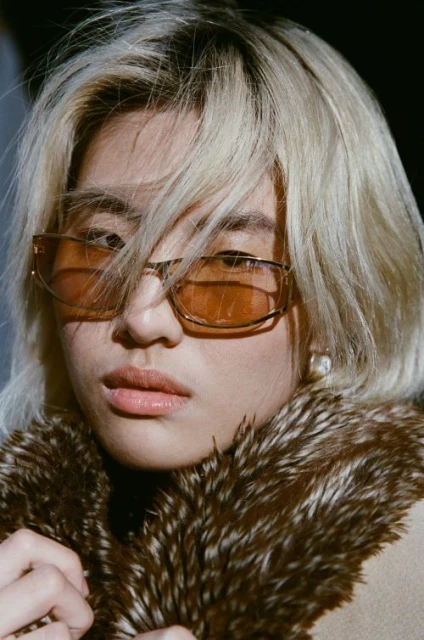 Close-up portrait of a young Asian woman wearing amber sunglasses and a fur-collared coat, gazing directly at the camera with a neutral expression.