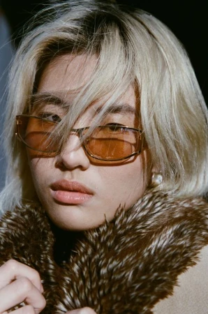 Close-up portrait of a young Asian woman wearing amber sunglasses and a fur-collared coat, gazing directly at the camera with a neutral expression.