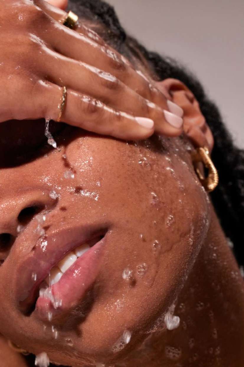 Close-up of a dark-skinned adult female washing her face with water, wearing gold earrings and rings. The background is neutral.