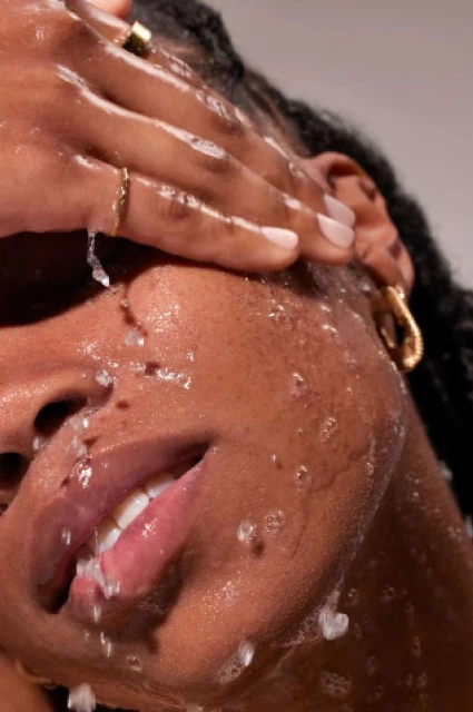 Close-up of a dark-skinned adult female washing her face with water, wearing gold earrings and rings. The background is neutral.