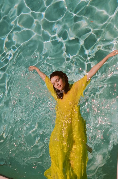 Woman in a yellow dress floats in a swimming pool. The sunlight creates beautiful reflections and patterns on the water's surface.