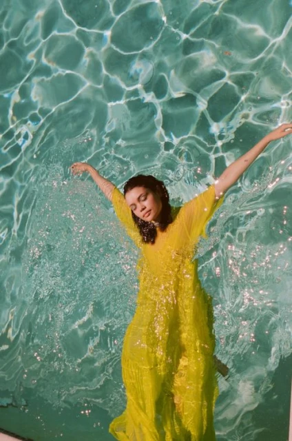 Woman in a yellow dress floats in a swimming pool. The sunlight creates beautiful reflections and patterns on the water's surface.