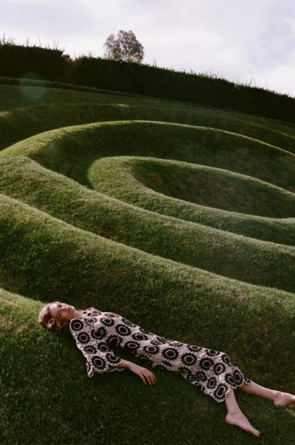 Woman in patterned dress lies on a grassy hillside. Green landscape of curved hills, relaxation, natural lighting. Fashion editorial.