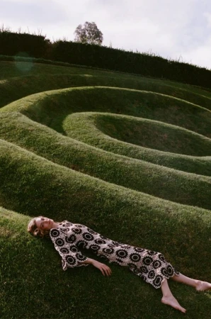 Woman in patterned dress lies on a grassy hillside. Green landscape of curved hills, relaxation, natural lighting. Fashion editorial.