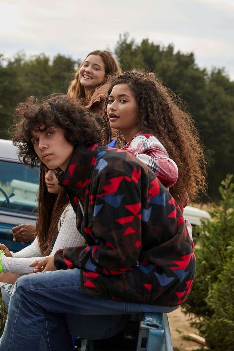 Group of young adults sit on vehicle in outdoor setting, trees and sky background. Casual fall fashion, close medium shot.
