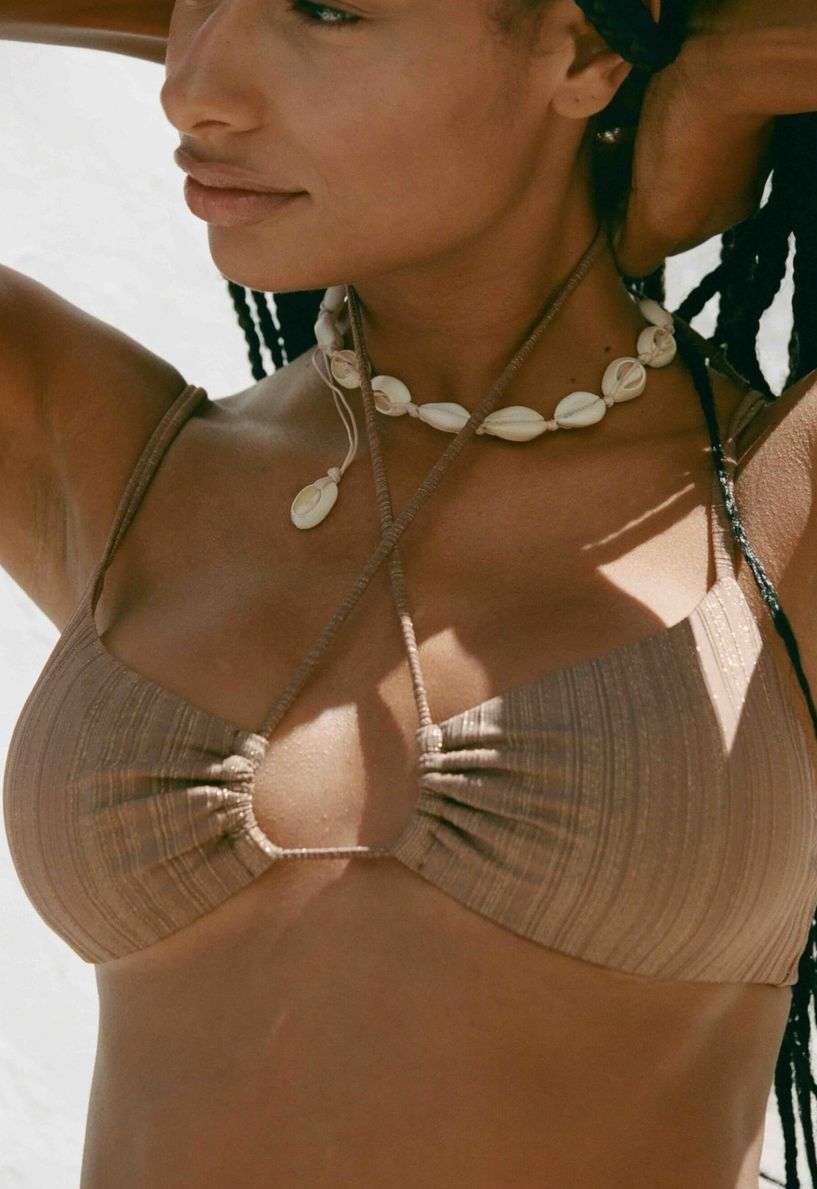 A tanned woman wears a brown bikini top and a shell necklace, posing against a blurred white background in an outdoor setting.