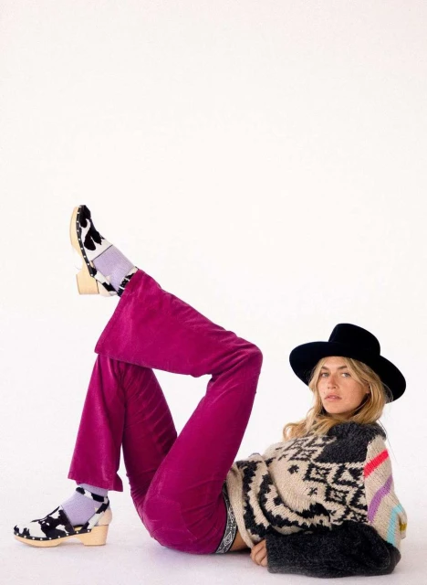 A woman poses lying down wearing a black hat, patterned sweater, purple velvet pants and socks and clogs with a cow print design.