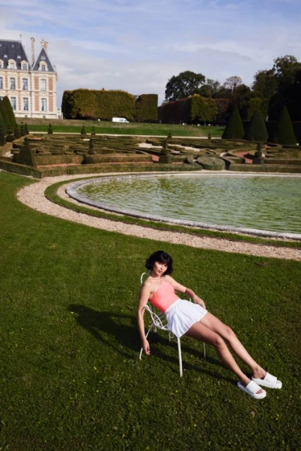 Woman in pink top and white skirt sits on a chair on the lawn in front of a pond and chateau under a blue sky with clouds.