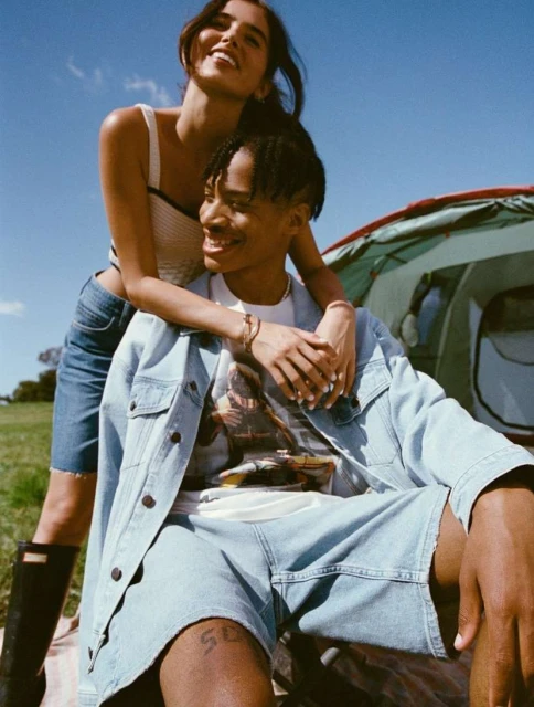 Outdoor fashion shot of a smiling couple, featuring an African American man wearing a denim jacket and a woman in a tank top and denim skirt.