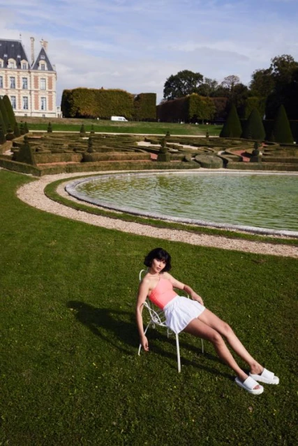 Woman in an orange top and white skirt reclines on a white chair on green grass in front of a pool in a garden with a building in the background.