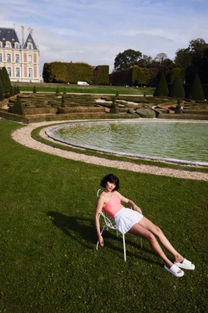 Woman in an orange top and white skirt reclines on a white chair on green grass in front of a pool in a garden with a building in the background.