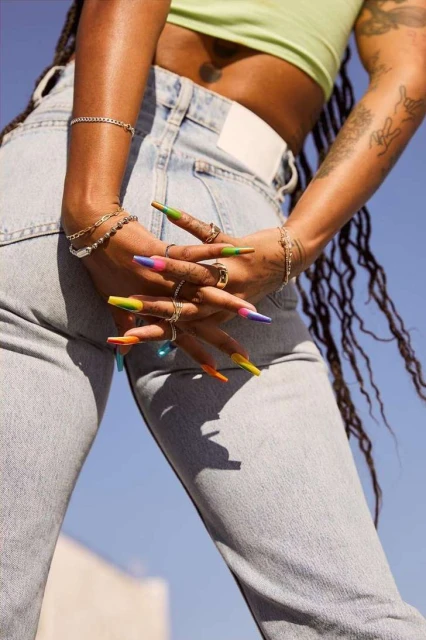 Close-up shot of an African American woman with long braids, light blue jeans, wearing colorful nails, bracelets, and rings against a blue sky.