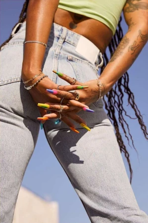 Close-up shot of an African American woman with long braids, light blue jeans, wearing colorful nails, bracelets, and rings against a blue sky.