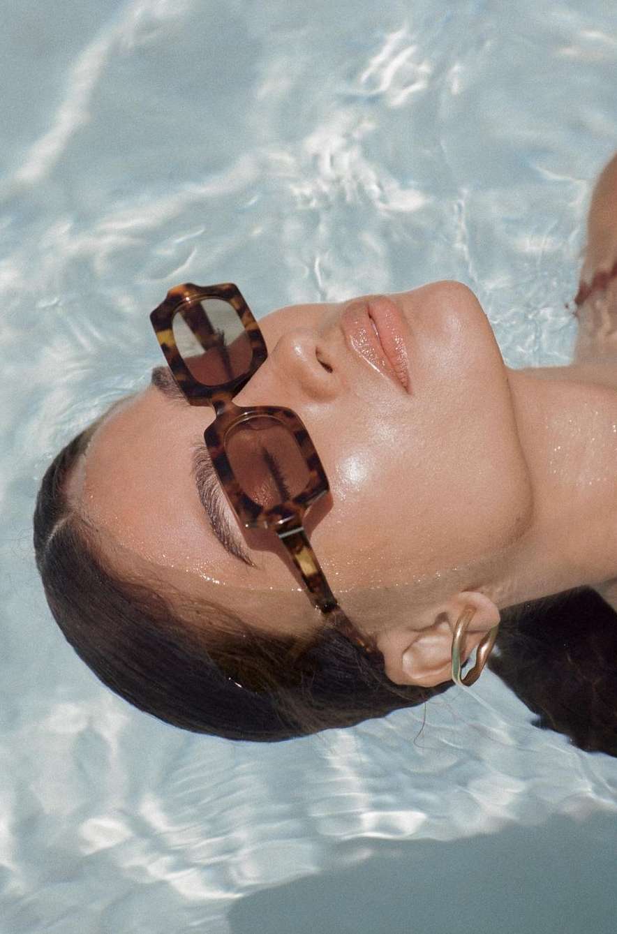 Close-up portrait of a woman wearing tortoiseshell sunglasses floating in clear swimming pool water by Ali Mitton.