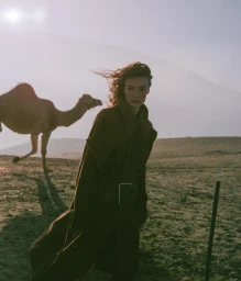 A woman in a long brown coat outdoors in a desert landscape with a camel standing in the background.