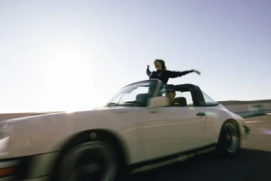 Woman standing with outstretched arms through the open top of a moving white sports car under a bright sky.