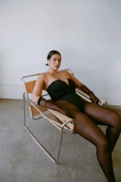 Woman in a black strapless bodysuit and sheer black gloves seated in a chrome and leather chair against a white wall