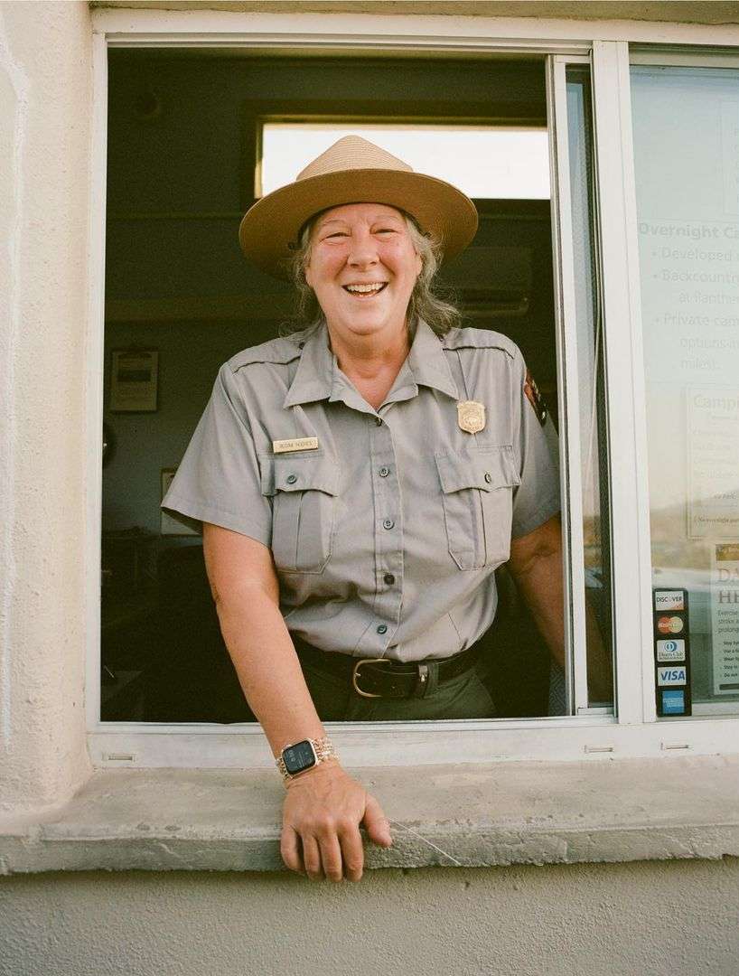 Smiling park ranger woman in uniform at booth, wearing a hat, with visible credit card logos. Shot in daylight with natural lighting.
