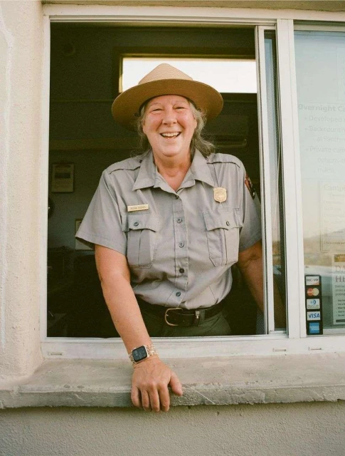 Smiling park ranger woman in uniform at booth, wearing a hat, with visible credit card logos. Shot in daylight with natural lighting.