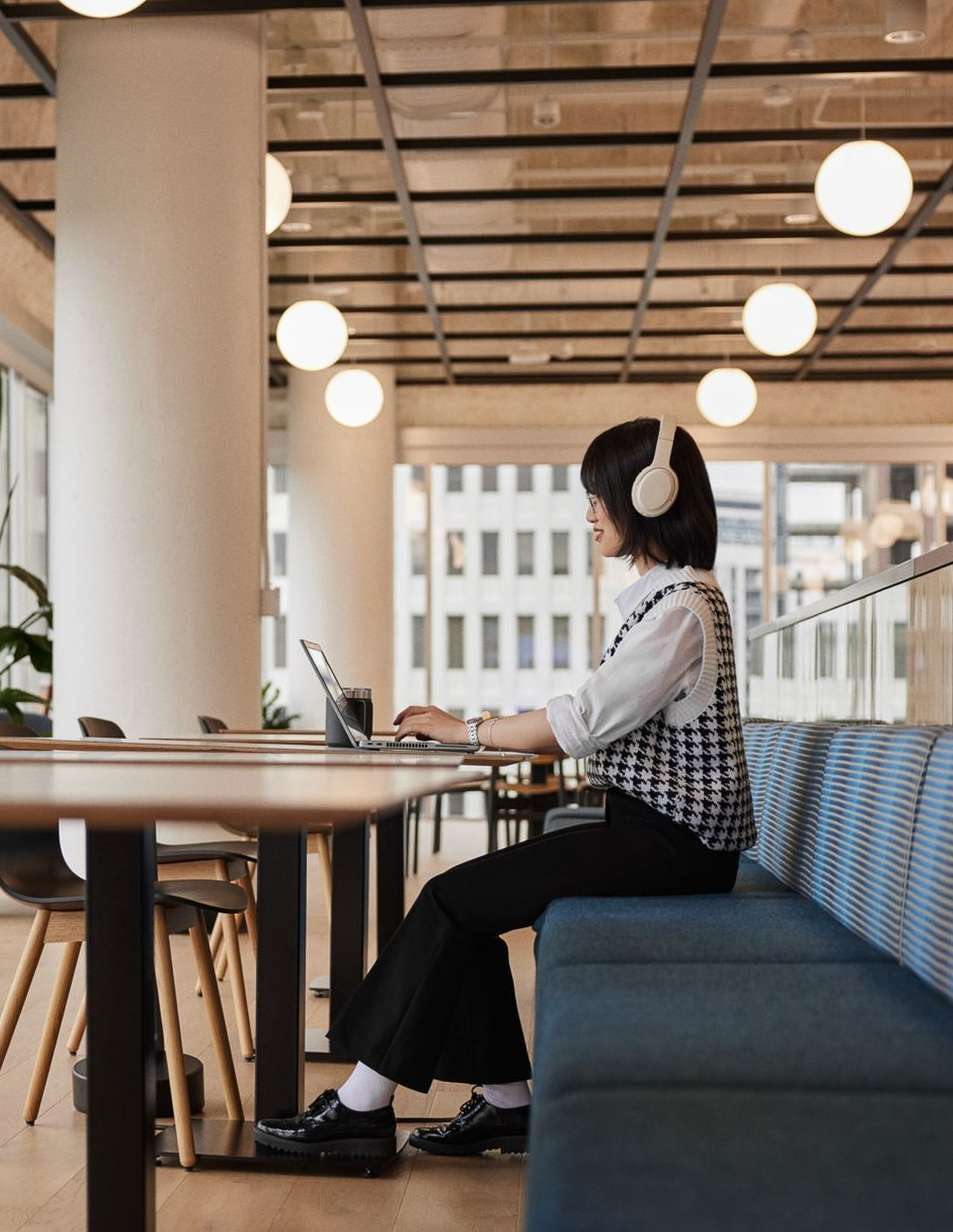 A woman wearing headphones is working on a laptop in a modern office, seated at a table with a blue bench, near a window overlooking the city.
