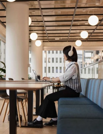A woman wearing headphones is working on a laptop in a modern office, seated at a table with a blue bench, near a window overlooking the city.