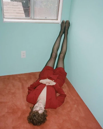 Woman lying on a red carpet with legs propped up on a teal wall, wearing a red suit and black tights in an indoor space with a window.