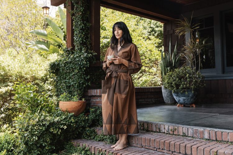 Woman in brown robe stands on brick steps with tea, surrounded by lush greenery and potted plants on a porch with a wooden exterior.