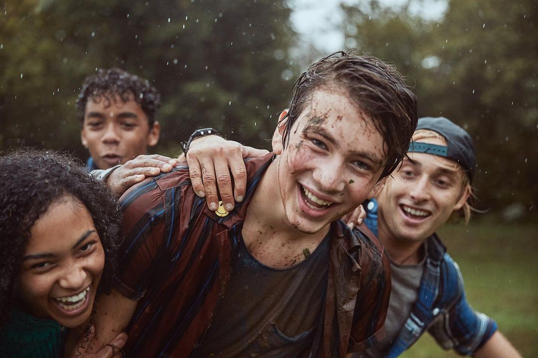 A group of four smiling teenagers stand together outdoors in the rain, covered in mud, with trees blurred in the background. 