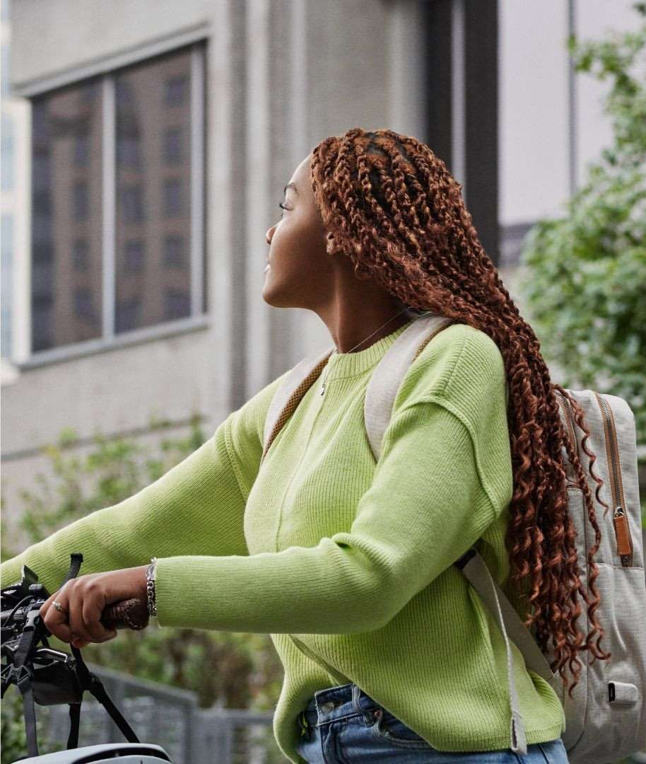 Woman with braided hair rides a bicycle outdoors in a city, wearing a green sweater and a backpack on a cloudy day.