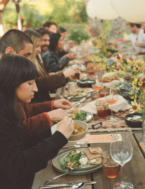 Outdoor banquet with a long wooden table laden with food and drinks surrounded by people at an outdoor daytime event.